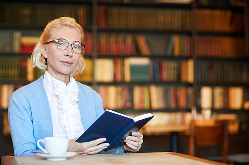 Aged blonde female with open book sitting in library or cafe at leisure and reading