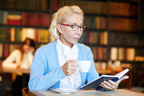 Aged woman with cup of tea or coffee reading curious novel in cozy cafe at leisure