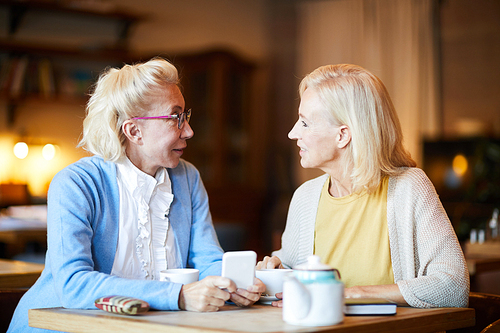 Two mature blonde women sitting by table in cafe and discussing online news by cup of tea