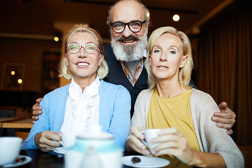 Senior bearded man embracing two blonde aged women during their hangout in cafe