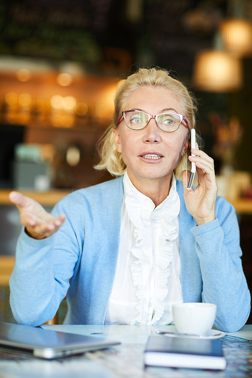 Busy aged female explaining something on smartphone while sitting by table and organizing work