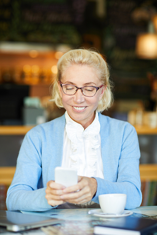 Happy woman texting in smartphone while sitting by table in cafe at coffee-break