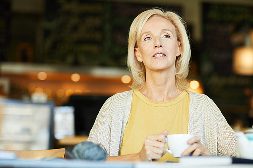 Pensive blonde aged woman with cup of tea or coffee sitting in cafe at leisure
