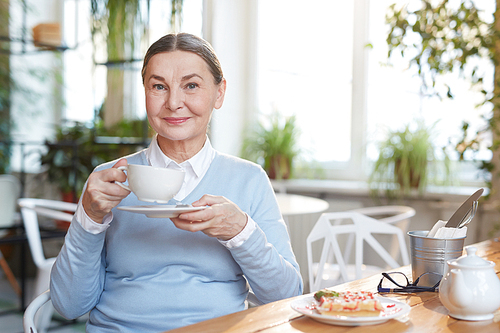 Smiling mature woman with cup of tea sitting by table and enjoying rest in cafe