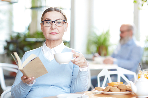 Elegant woman in eyeglasses and smart casual reading book while having tea with cookies in cafe