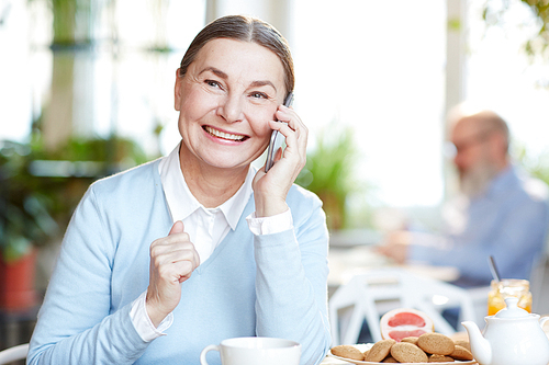 Cheerful mature woman with toothy smile talking by smartphone while having tea with cookies in cafe