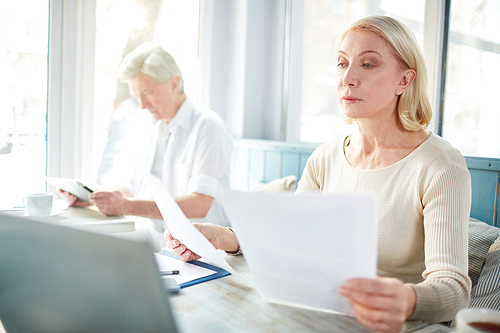 Mature blonde reading financial documents while working in cafe on background of colleague