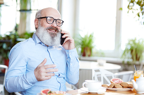 Senior man with grey beard sitting by table in cafe and talking on smartphone by cup of tea