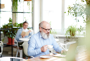 Senior man in blue shirt and eyeglassees having coffee and networking or talking through video-chat in cafe