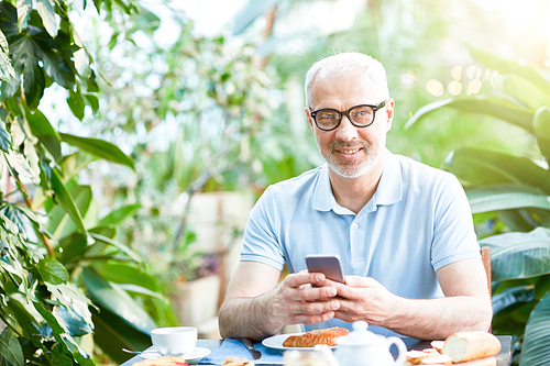 Happy man in eyeglasses  while texting in smartphone by served table during breakfast