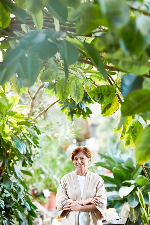 Casual aged female with her arms crossed on chest having promenade among green plants in the garden