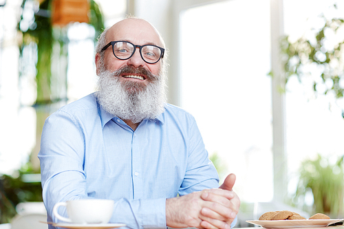 Happy aged man in eyeglasses spending time in cafe by cup of tea and cookies