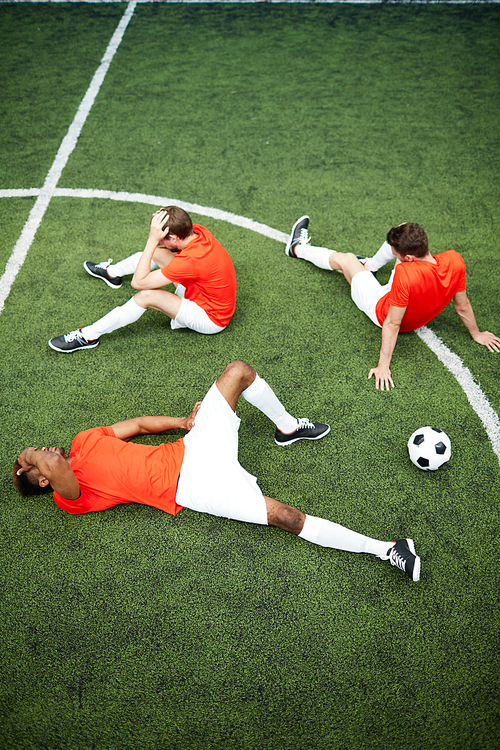 Three unlucky football players sitting and lying on green field after hard match or game