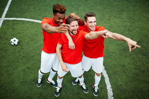 Three happy young footballers standing on green field, laughing and pointing forwards