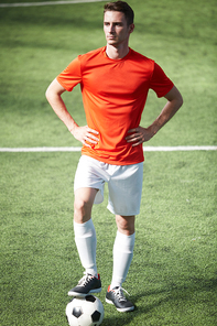 Young confident soccer player in akimbo pose standing on green field with one foot over ball