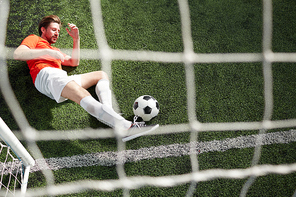 Top view of young goalkeeper catching soccer ball by leg while lying on green field by gates