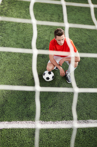 Young football player with ball squatting on green field in front of gates with net