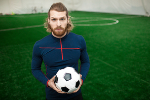 Serious young trainer of football team in activewear holding soccer ball while standing on pitch