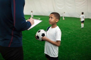 Adorable football player with ball talking to his trainer on pitch after game