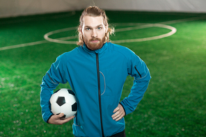 Young serious football player or trainer with ball standing on green pitch