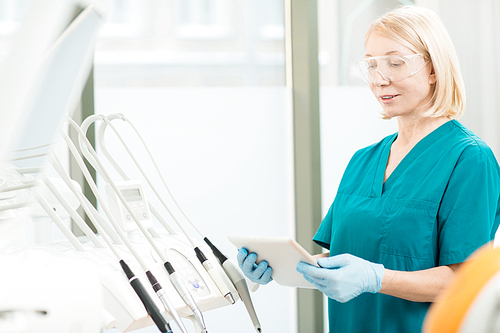 Mature professional dentist in protective eyeglasses, gloves and uniform using modern gadget to look for necessary information