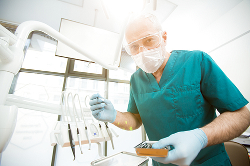 Contemporary dentist in protective mask, eyeglasses, gloves and uniform looking at patient before check-up