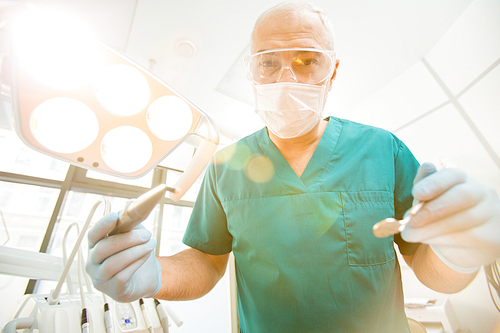 Mature dentist in uniform and protective mask and eyeglasses holding drill and mirror