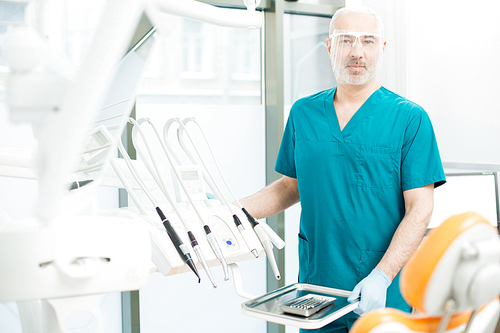 Confident dentist in unform and protective eyeglasses  by his workplace