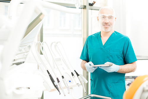 Confident dentist with small metallic tray standing by professional equipment in dental clinics