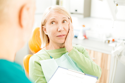 Mature woman keeping her hand on cheek while talking to dentist during appointment