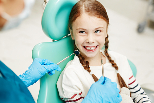 Cheerful excited pretty redhead girl with braids smiling at unrecognizable dentist with dental instruments in hands