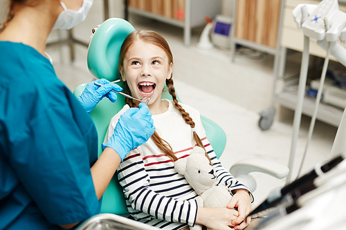 Positive cute girl with redhead braids holding toy while sitting in dental chair and keeping mouth open during dental checkup