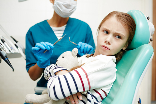 Gloomy frowning redhead girl embracing toy bear and turning away from dentist while sitting in dental chair and refusing to treat teeth