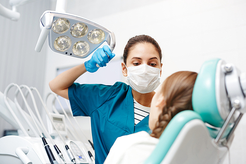 Serious confident young female pediatric dentist in mask and gloves adjusting lamp while holding checkup of childrens teeth