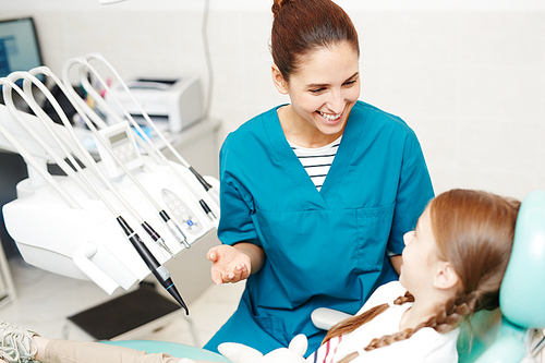 Cheerful excited young female dentist working with redhead girl: she gesturing while telling about dental health to kid