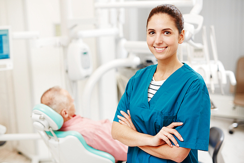 Cheerful confident successful young female dentist in uniform with crossed arms  while senior patient sitting in dental chair in background
