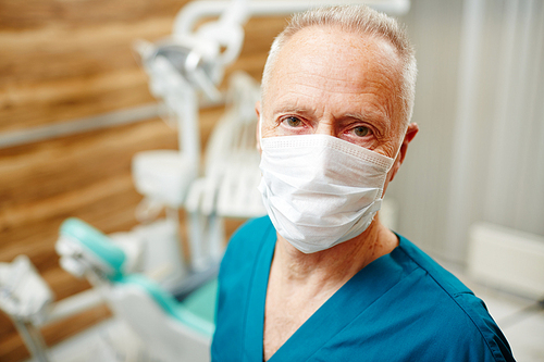 Portrait of serious confident handsome elderly male dentist with wrinkles wearing mask standing in own office