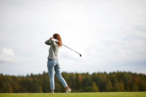 Young woman in activewear holding golf club after hitting ball while standing on the field