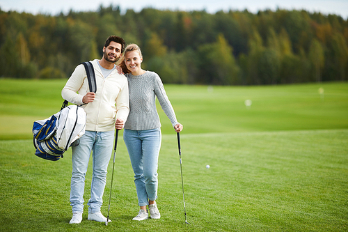 Happy young couple in casualwear standing on large green field for playing golf