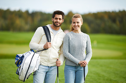 Couple of young golfers with equipment standing on large green field outdoors