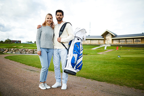 Young casual dates with equipment for playing golf standing on the road by green field