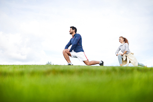 Young active man and woman in sportswear bending knees and stretching legs during outdoor workout