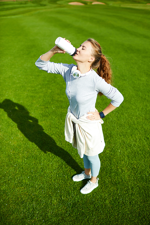 Active girl standing on green grass and drinking water from plastic bottle