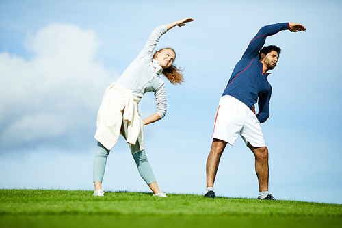 Happy young woman and her boyfriend in activewear side-bending against blue sky