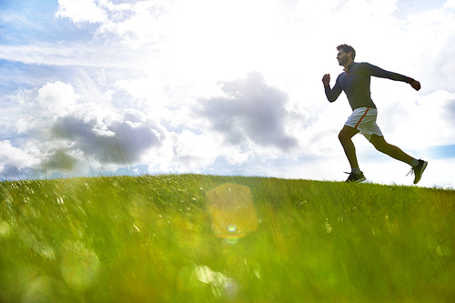 Young active man running down green field against cloudy sky on summer day