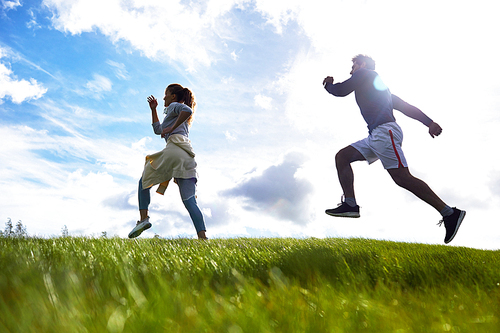 Active young couple running down green grass against cloudy sky on summer morning