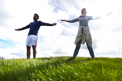 Young couple in activewear standing on green field with their arms outstretched