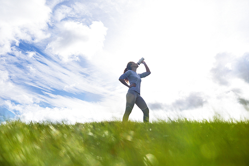 Thirsty sportswoman drinking water from bottle after workout while standing on large green field