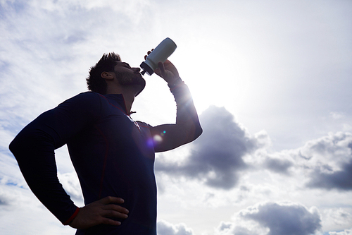 Active guy drinking water from plastic bottle against cloudy sky and sunshine