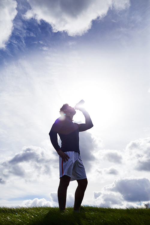Young man in activewear standing against sunshine on cloudy sky and having refreshment after workout